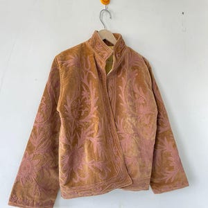 May include: A tan velvet jacket featuring an intricate, raised pink floral embroidery pattern across the front and sleeves. The jacket has a high collar, long sleeves, and a straight hemline, with a visible yellow interior lining at the neck.