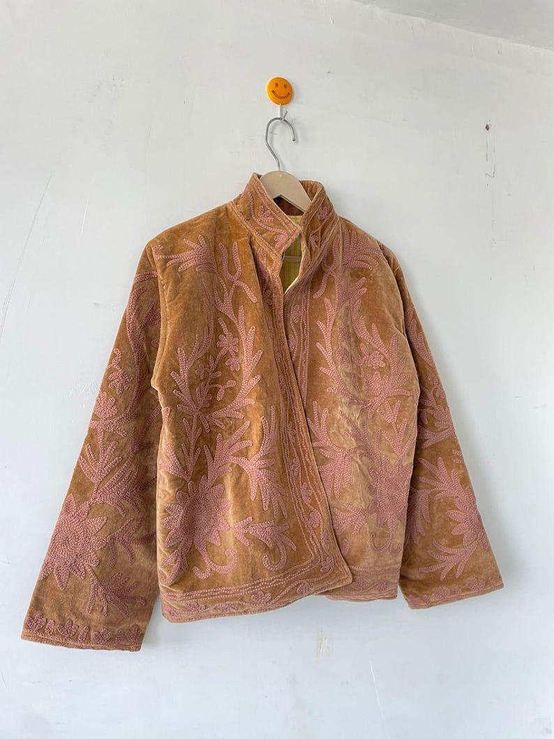 May include: A tan velvet jacket featuring an intricate, raised pink floral embroidery pattern across the front and sleeves. The jacket has a high collar, long sleeves, and a straight hemline, with a visible yellow interior lining at the neck.
