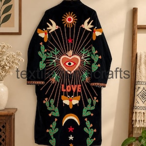 Puede incluir: Una bata negra de manga larga con bordados coloridos en la espalda. El diseño incluye un corazón central con un ojo, un sol, pájaros, polillas, cactus y una luna creciente. La palabra LOVE está bordada en letras rojas bajo el corazón.