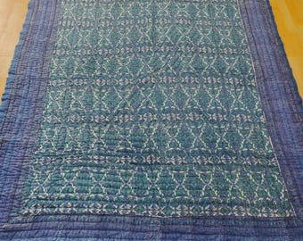 Könnte beinhalten: Eine gesteppte Tagesdecke in Blau- und Türkistönen. Die Mitte zeigt ein sich wiederholendes Blumenmuster in Türkis und Weiß, das von einem einfarbigen blauen Rand umgeben ist. Die Kanten sind mit einer dunkelblauen Einfassung versehen.