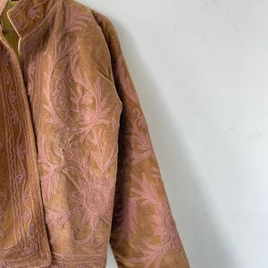 May include: A tan suede jacket featuring intricate pink floral embroidery across the front and sleeves. The jacket has a high collar, long sleeves, and a visible yellow and white striped lining on the inside of the front opening.