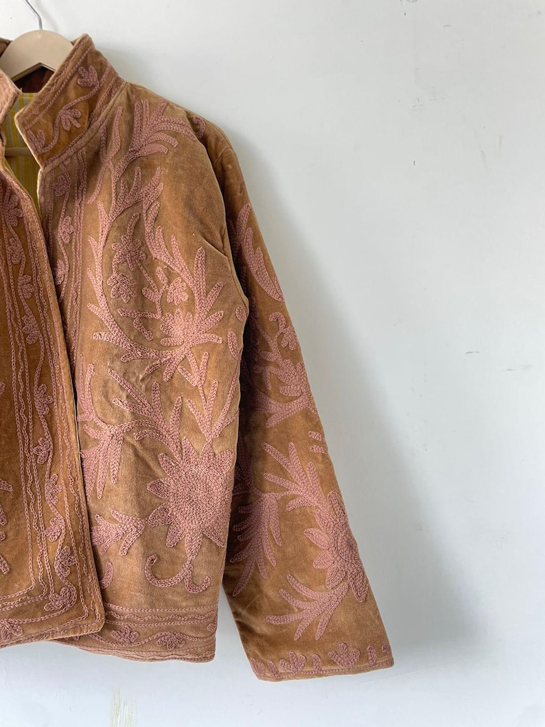 May include: A tan suede jacket featuring intricate pink floral embroidery across the front and sleeves. The jacket has a high collar, long sleeves, and a visible yellow and white striped lining on the inside of the front opening.