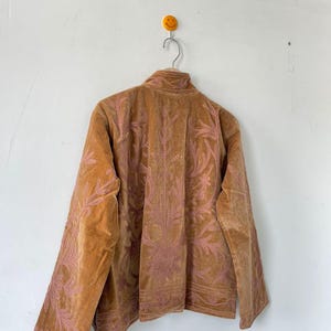 May include: A tan velvet jacket featuring intricate pink floral embroidery across the back and sleeves. The jacket has a high collar, long sleeves, and a structured fit, showcasing detailed stitching patterns that extend from the shoulders down.