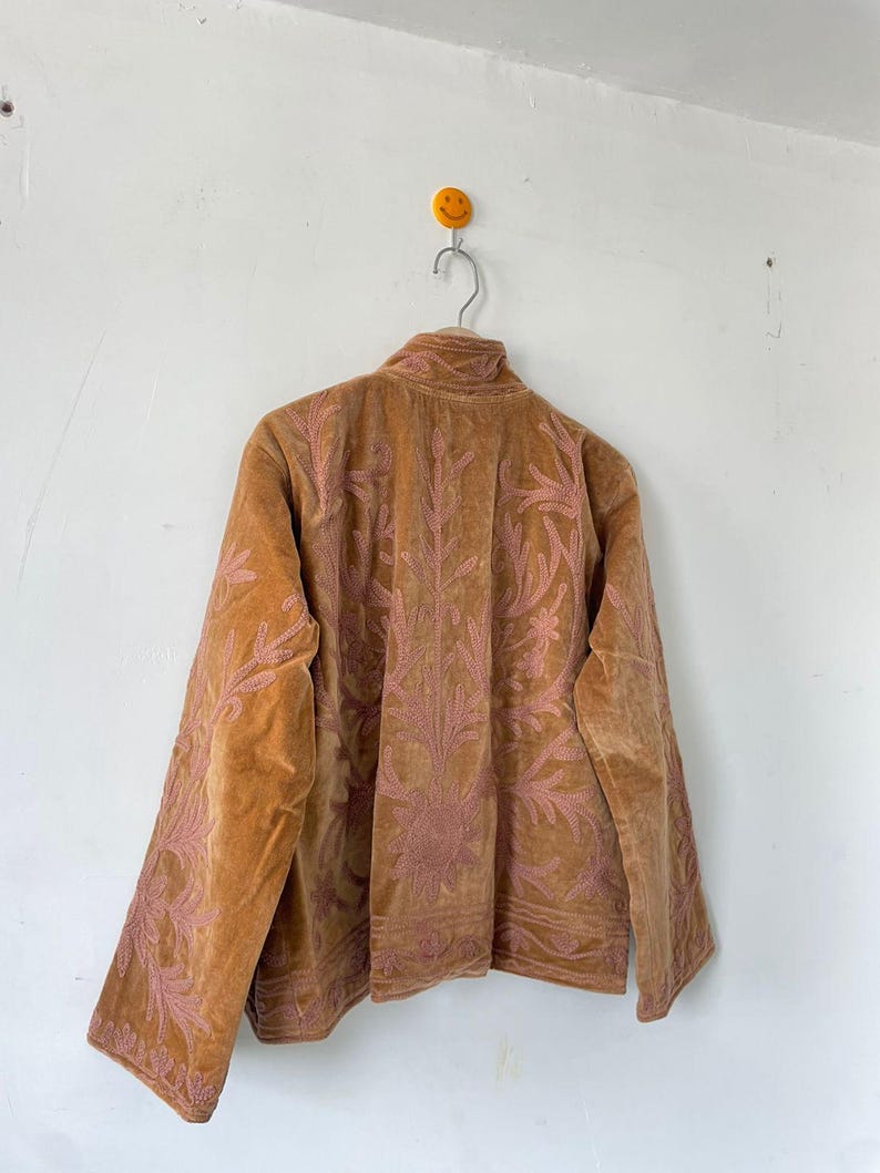 May include: A tan velvet jacket featuring intricate pink floral embroidery across the back and sleeves. The jacket has a high collar, long sleeves, and a structured fit, showcasing detailed stitching patterns that extend from the shoulders down.