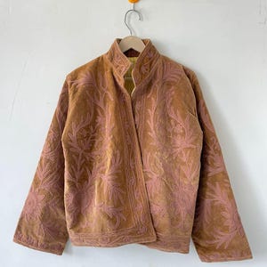 May include: A tan jacket featuring long sleeves, a high collar, and an open front design. The garment is decorated with intricate, light pink floral embroidery patterns that cover the front panels and extend across the sleeves and collar.