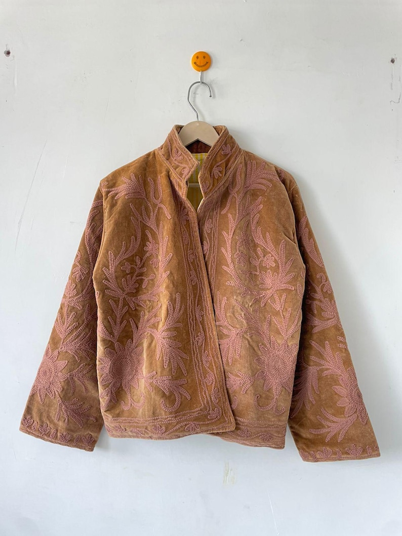 May include: A tan jacket featuring long sleeves, a high collar, and an open front design. The garment is decorated with intricate, light pink floral embroidery patterns that cover the front panels and extend across the sleeves and collar.