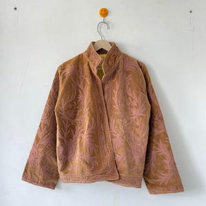 May include: A tan velvet jacket featuring an open front, a stand-up collar, and long sleeves. The garment is decorated with intricate, tonal pink floral embroidery patterns that cover the front panels and extend down the sleeves.