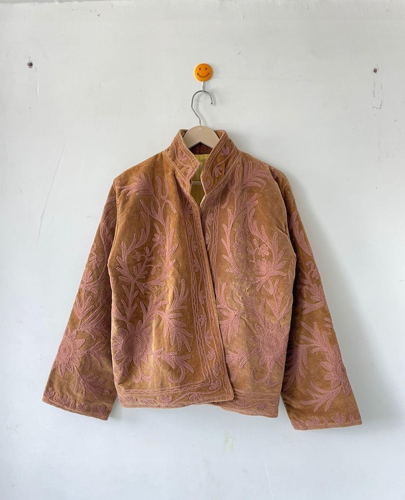 May include: A tan velvet jacket featuring an open front, a stand-up collar, and long sleeves. The garment is decorated with intricate, tonal pink floral embroidery patterns that cover the front panels and extend down the sleeves.