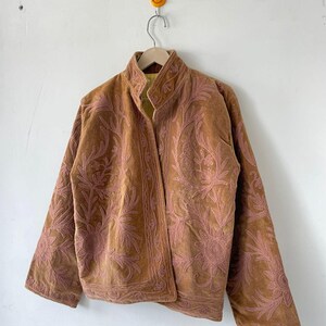 May include: A tan jacket featuring intricate pink floral embroidery across the front and sleeves. The garment has a high collar, long sleeves, and a straight hemline. The interior lining is visible at the collar and is a bright yellow color.
