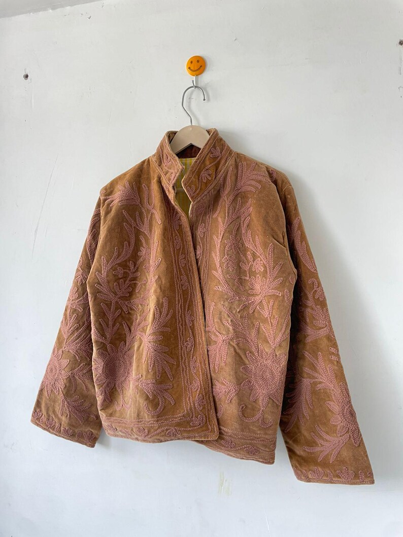 May include: A tan jacket featuring intricate pink floral embroidery across the front and sleeves. The garment has a high collar, long sleeves, and a straight hemline. The interior lining is visible at the collar and is a bright yellow color.