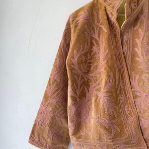 May include: A tan velvet jacket featuring intricate pink floral embroidery across the front and sleeves. The garment has a high collar, long sleeves, and a structured open front design, showcasing detailed stitching patterns throughout the fabric.