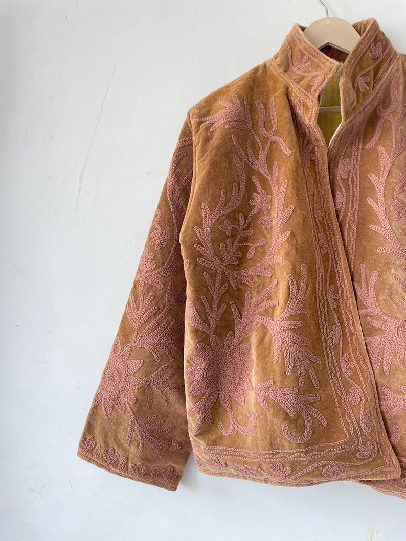 May include: A tan velvet jacket featuring intricate pink floral embroidery across the front and sleeves. The garment has a high collar, long sleeves, and a structured open front design, showcasing detailed stitching patterns throughout the fabric.