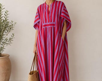 May include: A floor-length kaftan dress featuring vertical stripes in vibrant red and purple. The garment has a split V-neckline, wide short sleeves, and a gathered waist detail. It is paired with a woven straw tote bag and brown leather sandals.