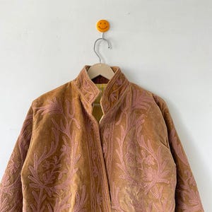 May include: A tan velvet jacket featuring an intricate, raised pink floral embroidery pattern across the front panels and collar. The jacket has a high neckline and a visible yellow and white striped lining visible at the inner collar area.