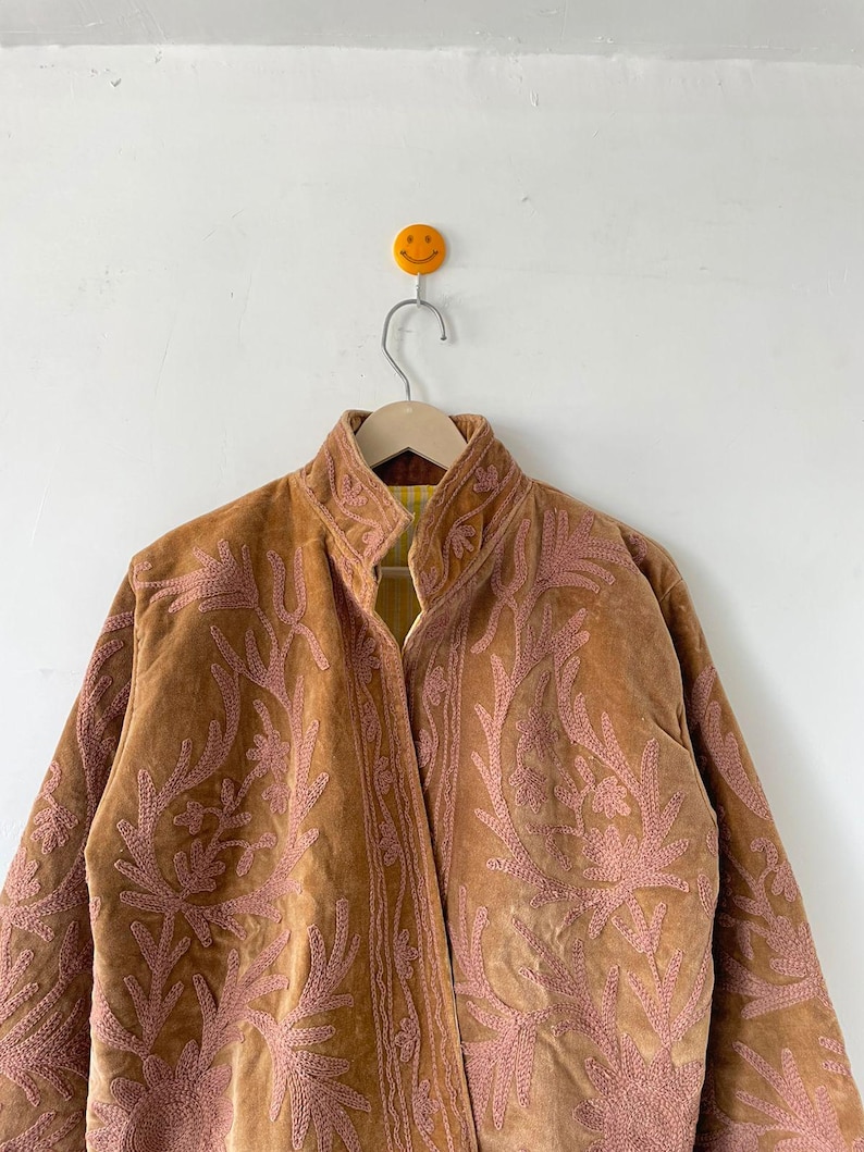 May include: A tan velvet jacket featuring an intricate, raised pink floral embroidery pattern across the front panels and collar. The jacket has a high neckline and a visible yellow and white striped lining visible at the inner collar area.