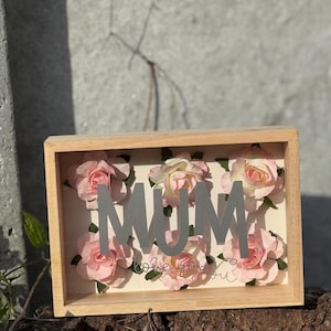 Op de afbeelding: Een houten schaduwdoos met het woord "MUM" in zilveren letters, omringd door roze rozen. De zin "love you" staat in sierlijke letters onder de bloemen. De doos is lichtbruin.