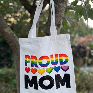 Op de afbeelding: Witte canvas draagtas met de tekst "PROUD MOM" in regenboogkleuren en zwart. Het ontwerp bevat regenboogkleurige harten. De tas heeft twee witte handvatten en hangt aan een tak.