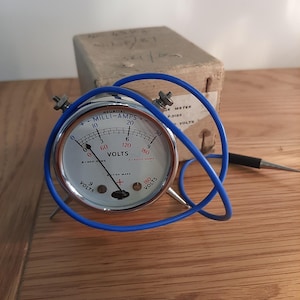 May include: A vintage electrical meter with a white face, black markings, and a blue wire. The meter reads "Milli-Amps" and "Volts." The device is housed in a silver casing and has a black probe. The meter is on a wooden surface.