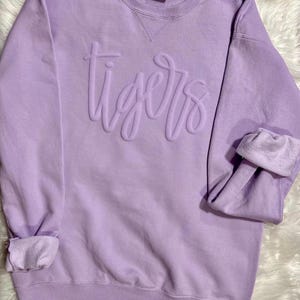 May include: A light purple sweatshirt with the word "tigers" in raised lettering across the chest. The sleeves are cuffed, and the garment has a crew neck. The sweatshirt is made of a soft material.