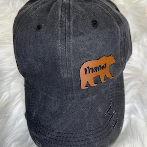 May include: Distressed black baseball cap with a brown leather patch featuring a bear silhouette and the word "Mama" in script. The cap has a pre-curved bill and distressed detailing.
