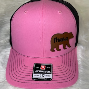 May include: Pink trucker hat with a black mesh back. The hat features a brown bear graphic with the word "Mama" and a Richardson tag. The hat is adjustable.