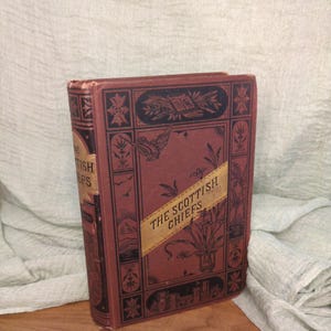 May include: Antique book titled "The Scottish Chiefs" with a maroon cover and gold lettering. The cover features black decorative elements and a gold banner. The book is standing upright on a wooden surface.