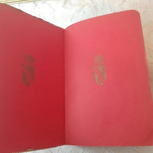 May include: Open book with red pages. Each page features a gold emblem in the upper center. The emblem is a shield shape with a diagonal stripe and a crown at the top. The book is open on a white lace surface.