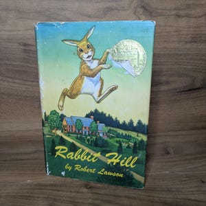 May include: A hardcover book titled Rabbit Hill by Robert Lawson, featuring a brown rabbit illustration, a house in a green landscape, and a gold Newbery Medal sticker on the front cover. The cover art displays a blue sky and green trees.