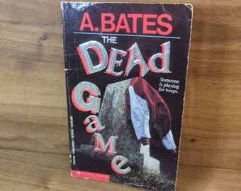Puede incluir: Un libro de bolsillo titulado The Dead Game de A. Bates con una portada negra que muestra una lápida y una chaqueta universitaria de color granate y blanco colgada sobre ella. El título está en letras grandes plateadas y rojas, y el texto Someone is playing for keeps aparece a la derecha.