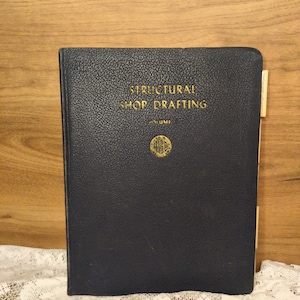 May include: A vintage navy blue book titled "STRUCTURAL SHOP DRAFTING" with gold lettering. The book has a textured cover and a gold emblem. A tan bookmark is visible on the right side of the book.