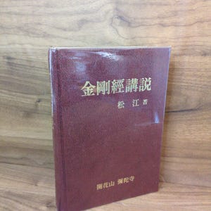 May include: A maroon book with Japanese text on the cover. The book has a green ribbon bookmark and is on a wooden surface. The text on the cover is in gold lettering.