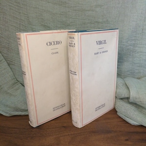 May include: Two vintage books with off-white covers. One book is titled "Cicero" and the other "Virgil." Both books have red and blue accents and the words "Interlinear Translation" printed on the spine. The books are standing upright on a wooden surface.