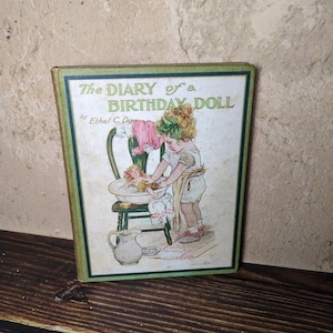 May include: A hardcover book titled The Diary of a Birthday Doll by Ethel C. Dow with a green border. The cover illustration depicts a child with a green hair bow bathing a doll in a basin on a chair, with a pitcher on the floor nearby.