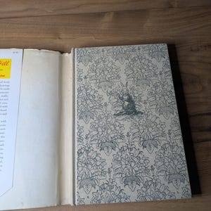 May include: A hardcover book with a light gray cover featuring a repeating black line-art pattern of flowers and leaves, with a central illustration of a rabbit. The book has a white dust jacket flap with visible text and a yellow rectangular label.