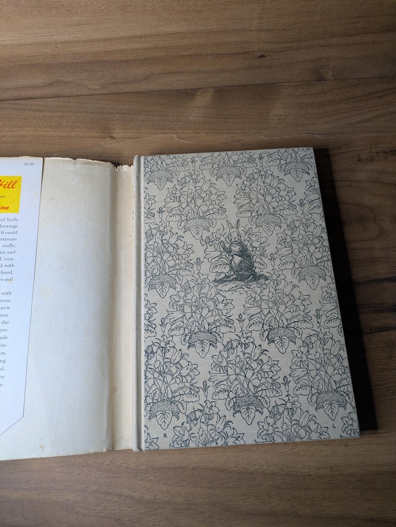 May include: A hardcover book with a light gray cover featuring a repeating black line-art pattern of flowers and leaves, with a central illustration of a rabbit. The book has a white dust jacket flap with visible text and a yellow rectangular label.