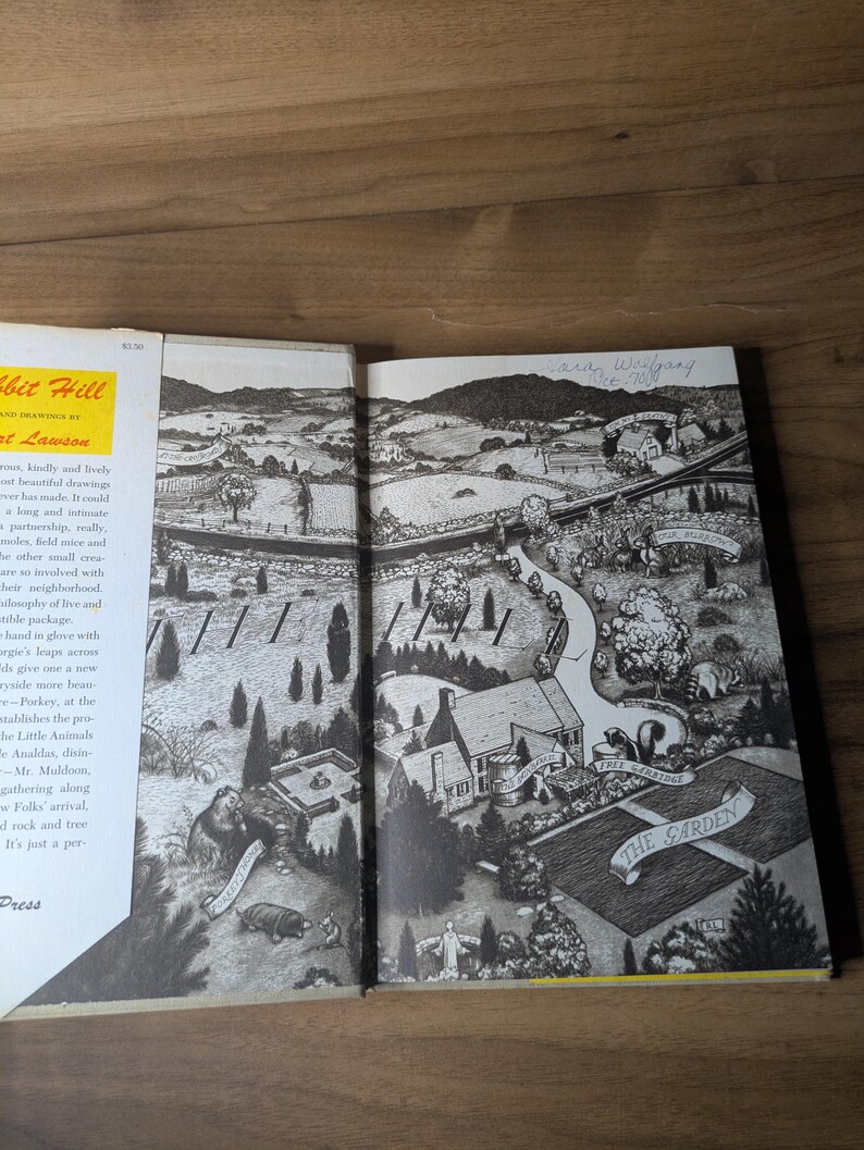 May include: An open hardcover book featuring a black and white ink illustration of a rural landscape with rolling hills, a house, and a garden. The left page shows text about Rabbit Hill by Robert Lawson, while the right page displays a detailed map.