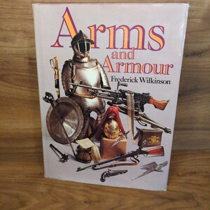 May include: A book titled "Arms and Armour" by Frederick Wilkinson. The cover features a knight in shining armor, a machine gun, and various historical weapons. The title is in purple and orange.