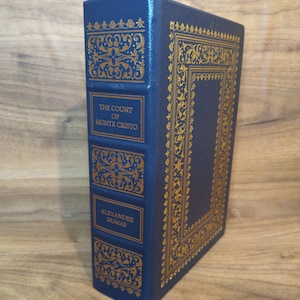 May include: A hardcover book titled "The Count of Monte Cristo" by Alexandre Dumas. The book has a dark blue cover with gold decorative accents and lettering. The book is standing upright on a wooden surface.