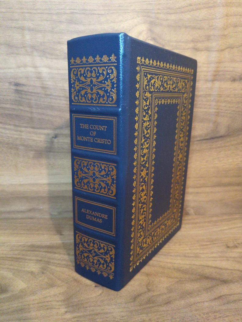 May include: A hardcover book titled "The Count of Monte Cristo" by Alexandre Dumas. The book has a dark blue cover with gold decorative accents and lettering. The book is standing upright on a wooden surface.