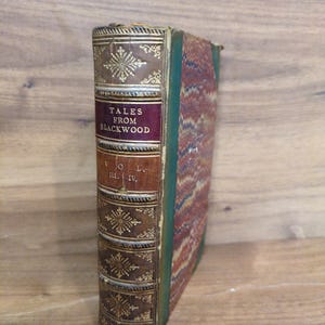 May include: A hardcover book titled Tales from Blackwood Vol. III. IV. featuring a brown leather spine with gold embossed floral patterns and lettering, and a marbled cover with green, red, and blue patterns on the side and back panels.