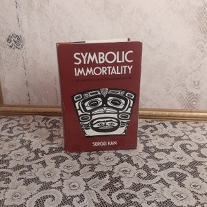 May include: A hardcover book titled "Symbolic Immortality" by Sergei Kan. The book has a maroon cover with white text and a black and white graphic. The subtitle reads "The Tlingit Potlatch of the Nineteenth Century."