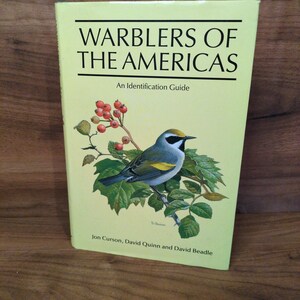 May include: A book titled "Warblers of the Americas" with a light yellow cover. The cover features a detailed illustration of a colorful bird perched on a leafy branch with red berries. The book is an identification guide.