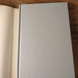 May include: A hardback book with a pale grey cover and a textured surface. The book is open, revealing the off-white pages. The spine is a darker grey. The book is on a wooden surface.