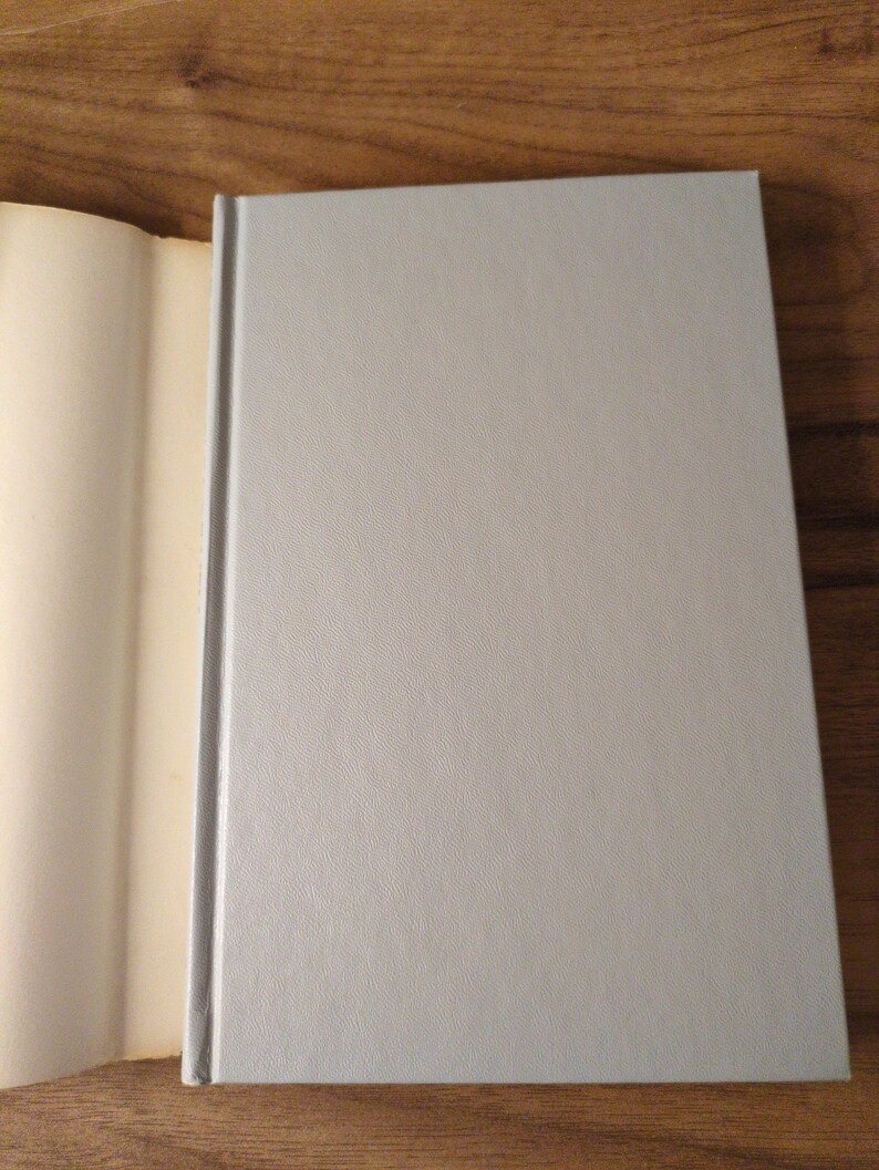 May include: A hardback book with a pale grey cover and a textured surface. The book is open, revealing the off-white pages. The spine is a darker grey. The book is on a wooden surface.