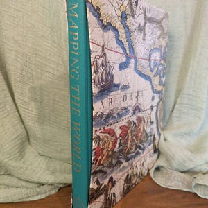 May include: A hardcover book titled "Mapping the World" with a teal spine and gold lettering. The cover features a vintage map design with illustrations of ships and figures, in shades of blue, beige, and red. The book is displayed upright.
