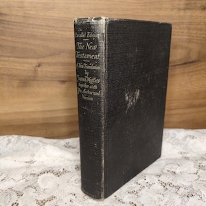 May include: A vintage black hardcover book titled "The New Testament" by James Moffatt. The book is a parallel edition with a new translation and the Authorized Version. The book is standing upright on a white lace surface.
