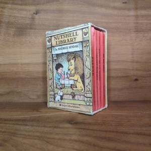 May include: A vintage Nutshell Library book set by Maurice Sendak. The cover features a colorful illustration of a boy and a lion at a table. The set includes four small books with red spines, displayed on a wooden surface.