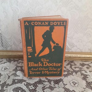 K&ouml;nnte beinhalten: Vintage-Buch mit dem Titel "The Black Doctor And Other Tales of Terror & Mystery" von A. Conan Doyle. Das Cover ist orange mit einer gr&uuml;nen Silhouette einer Person, die eine Tasche tr&auml;gt. Das Buch liegt auf einer wei&szlig;en Spitzentischdecke.