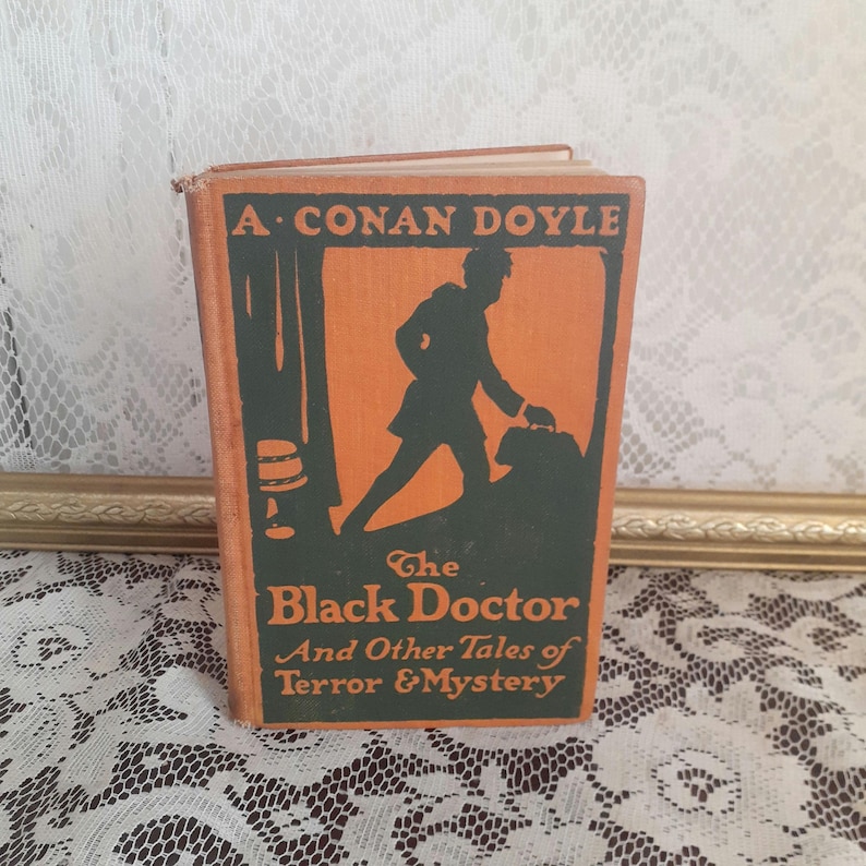 K&ouml;nnte beinhalten: Vintage-Buch mit dem Titel "The Black Doctor And Other Tales of Terror & Mystery" von A. Conan Doyle. Das Cover ist orange mit einer gr&uuml;nen Silhouette einer Person, die eine Tasche tr&auml;gt. Das Buch liegt auf einer wei&szlig;en Spitzentischdecke.