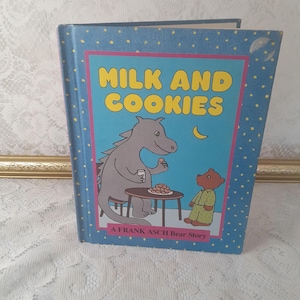 May include: A blue book with white polka dots on the cover. The title is "Milk and Cookies" in white letters. There is a picture of a gray dragon and a brown bear on the cover. The text at the bottom of the cover reads "A Frank Asch Bear Story".